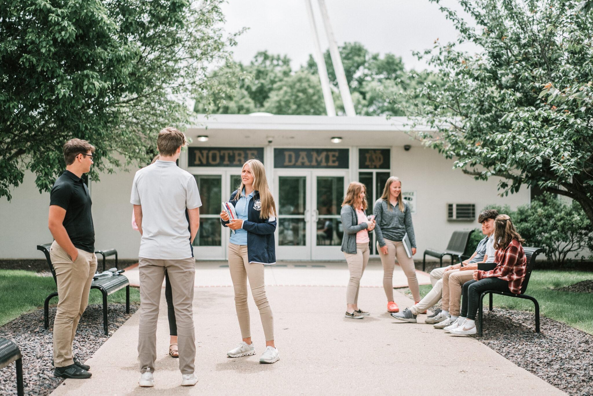 Students at Quincy Notre Dame High School socializing in front of the school entrance on a tree-lined walkway.