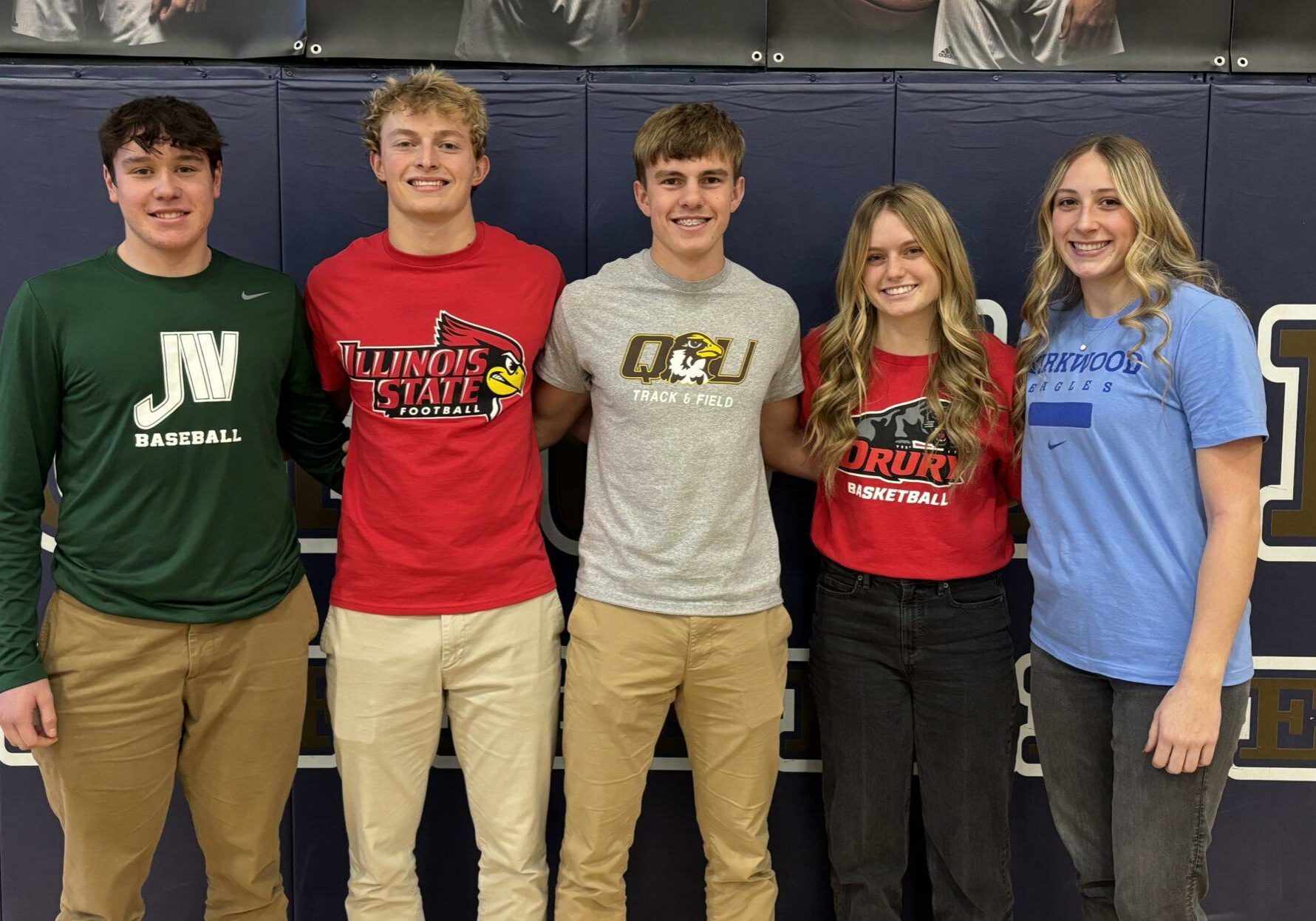 Five Quincy Notre Dame High School student-athletes posing together on signing day, wearing college shirts representing their future sports teams.