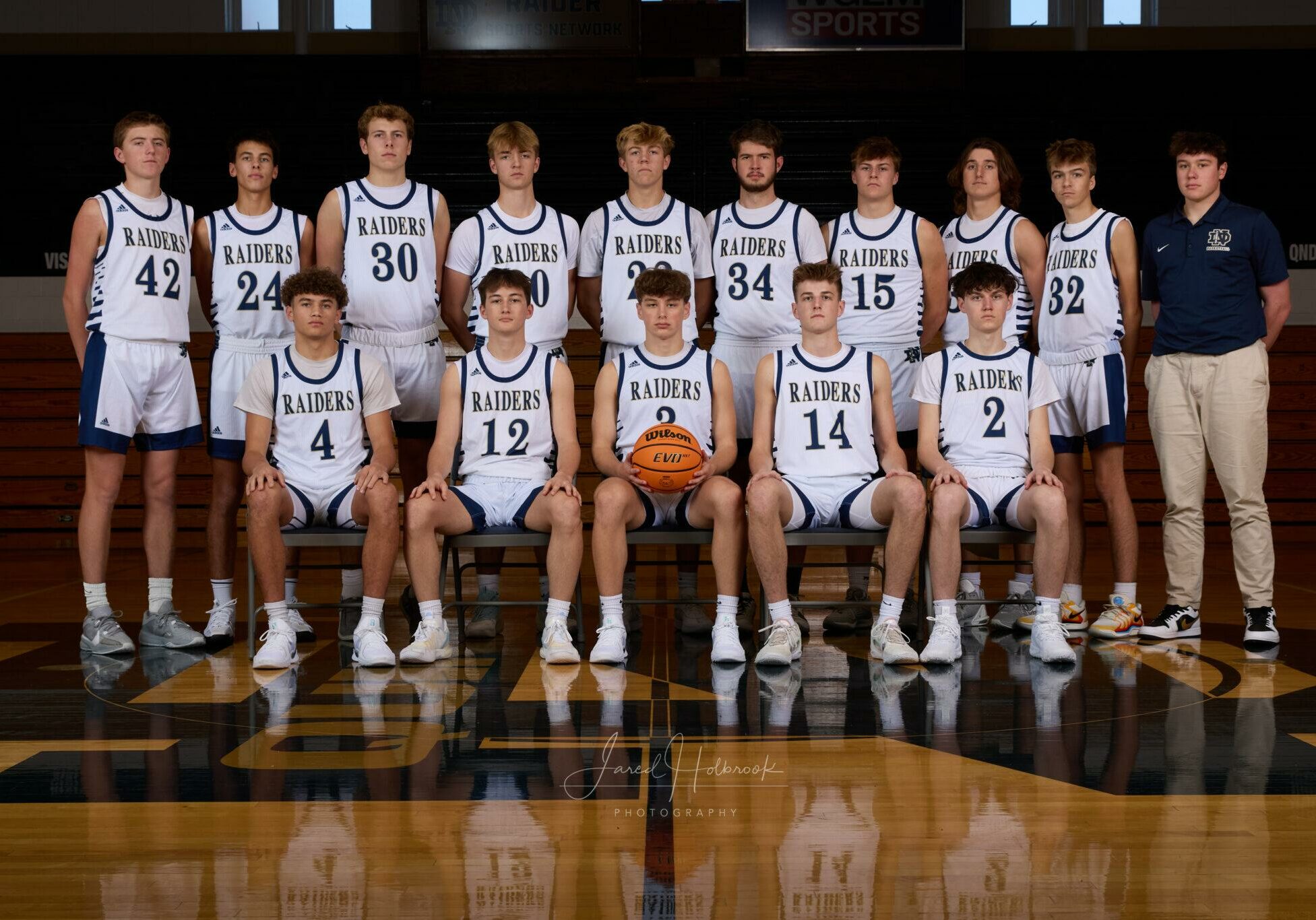 2024-25 Quincy Notre Dame varsity boys basketball team posing for a team photo in uniform on the gym court.