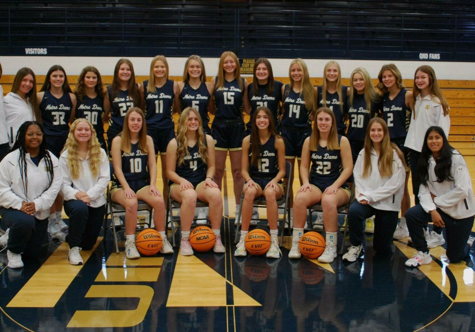 2024-25 Quincy Notre Dame varsity girls basketball team posing in uniform on the gym court.