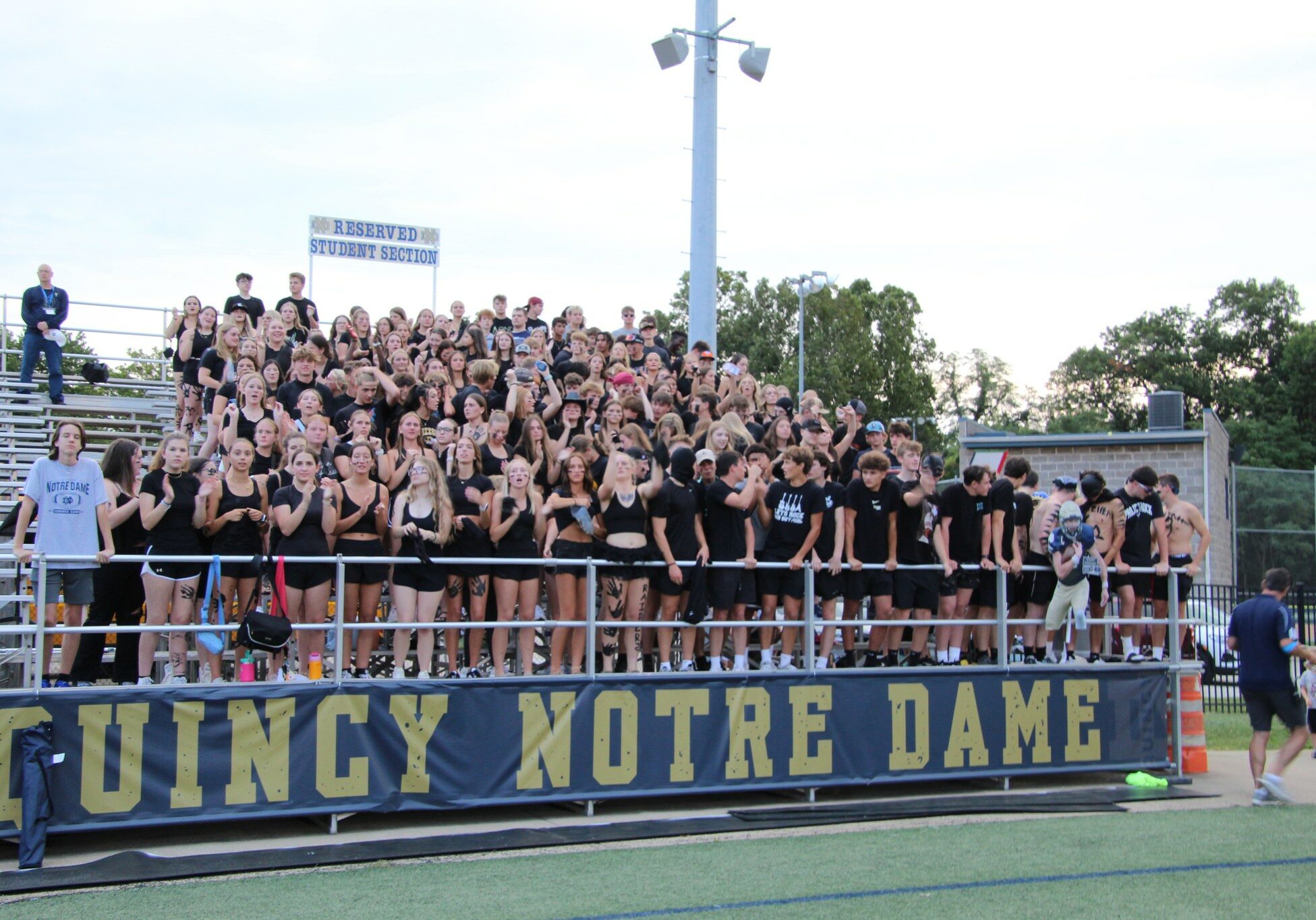 Quincy Notre Dame High School student section dressed in black, cheering from the bleachers during a school sports event.
