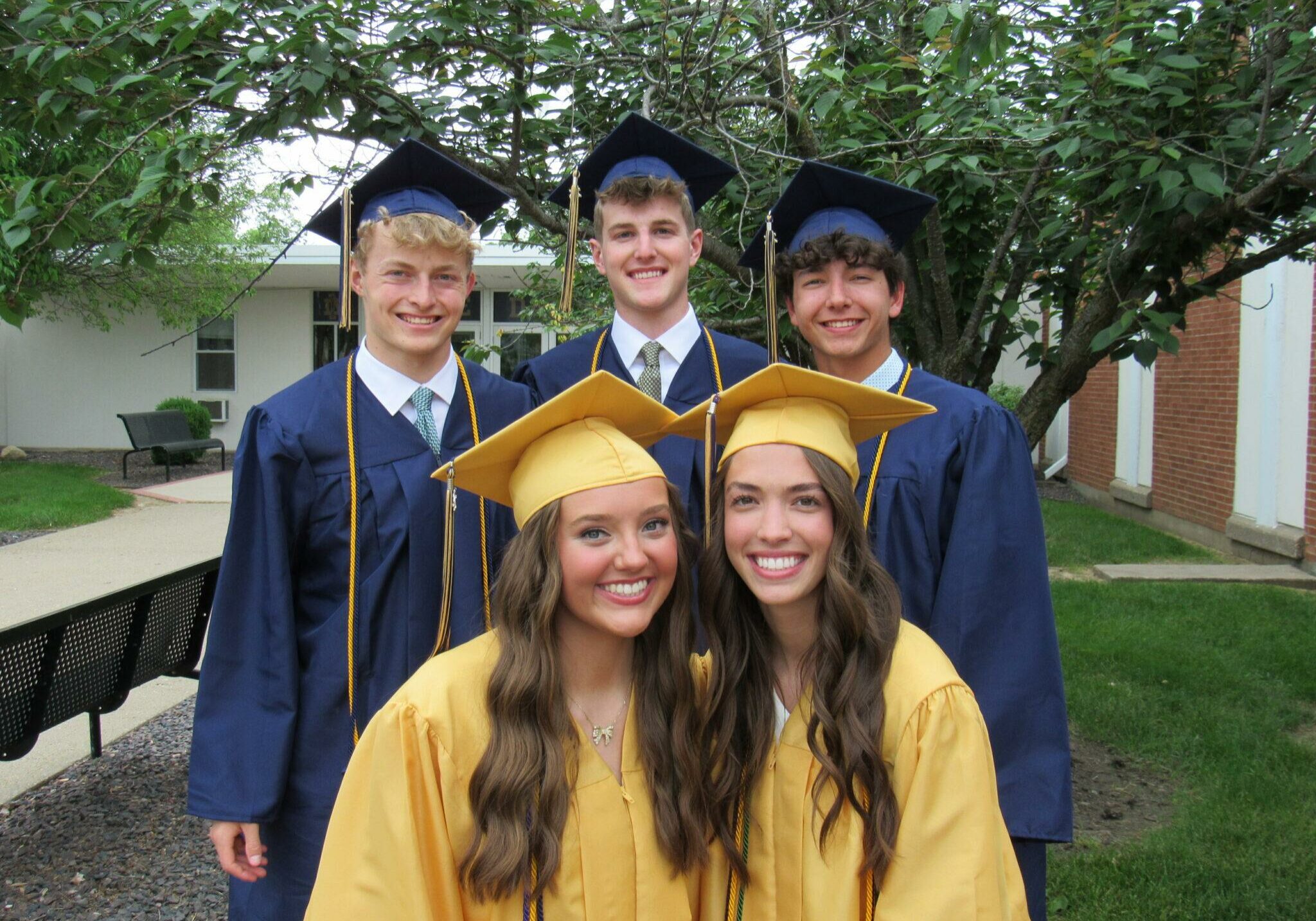 Five Quincy Notre Dame High School graduates posing outdoors in caps and gowns—three in navy blue and two in gold—smiling proudly on graduation day.