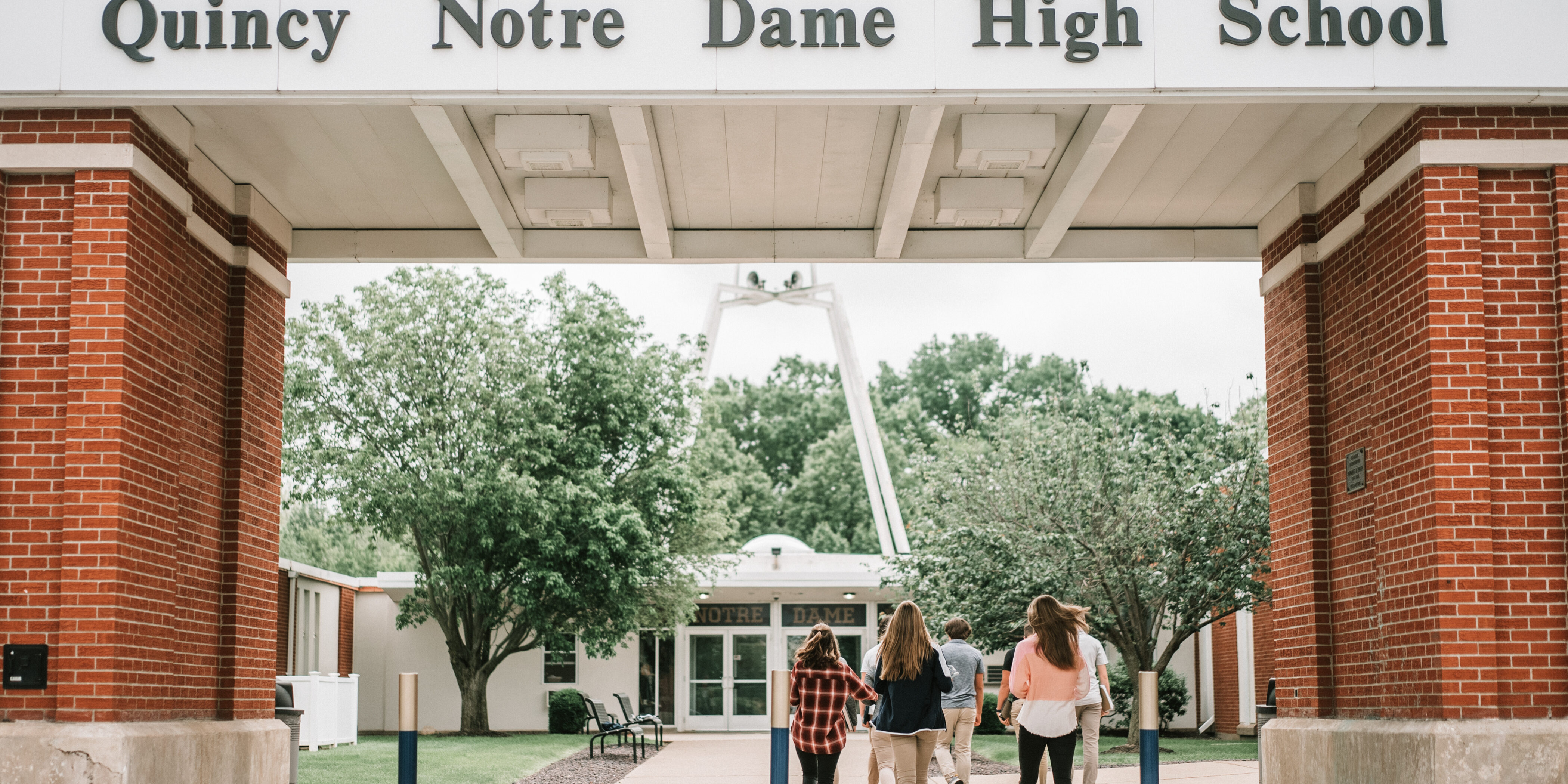 Students walking toward the entrance of Quincy Notre Dame High School under a large brick and white archway displaying the school's name.