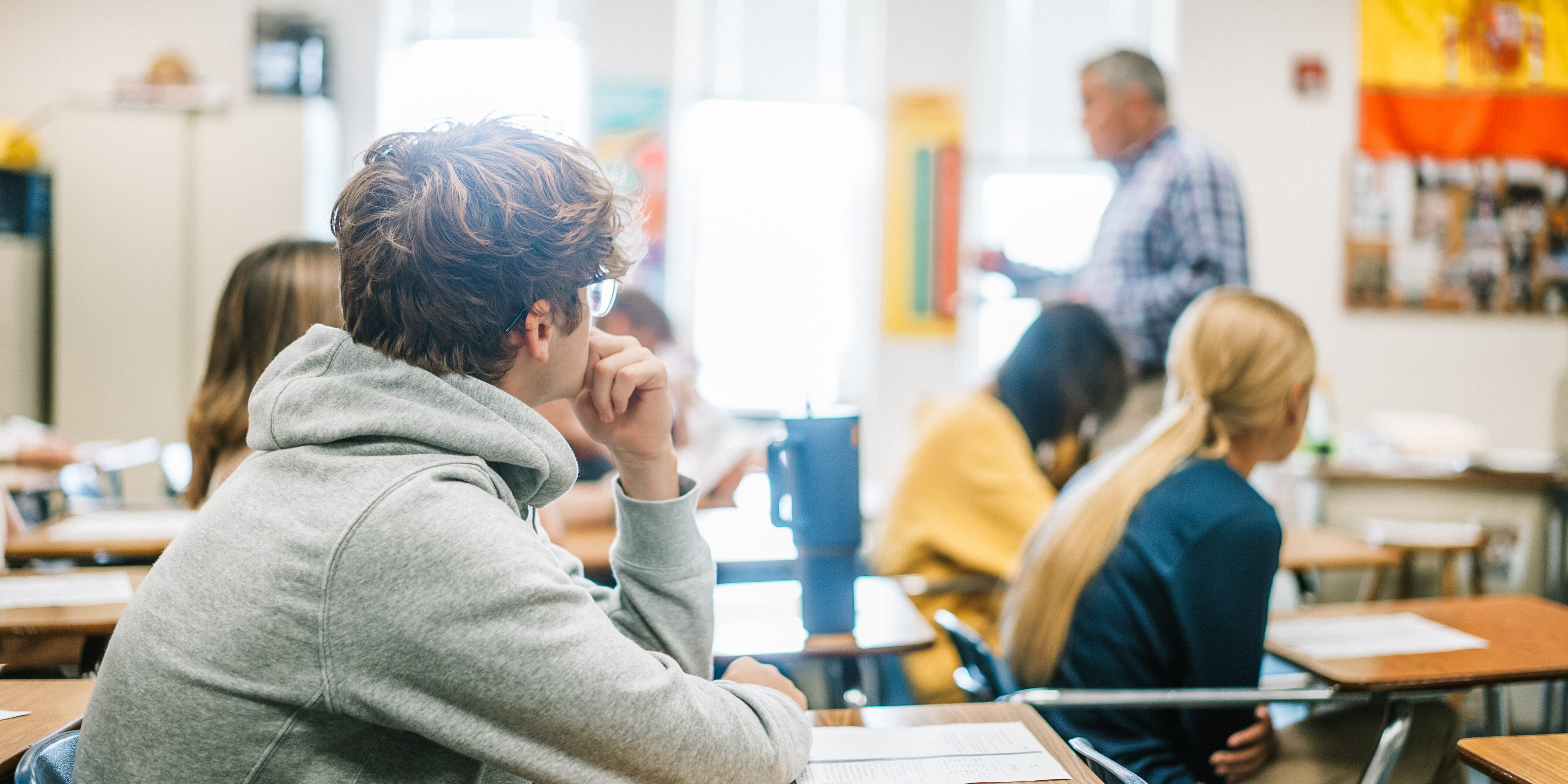 High school students seated at desks in a classroom attentively listening to a teacher speaking at the front of the room, with a Spanish flag hanging on the wall.
