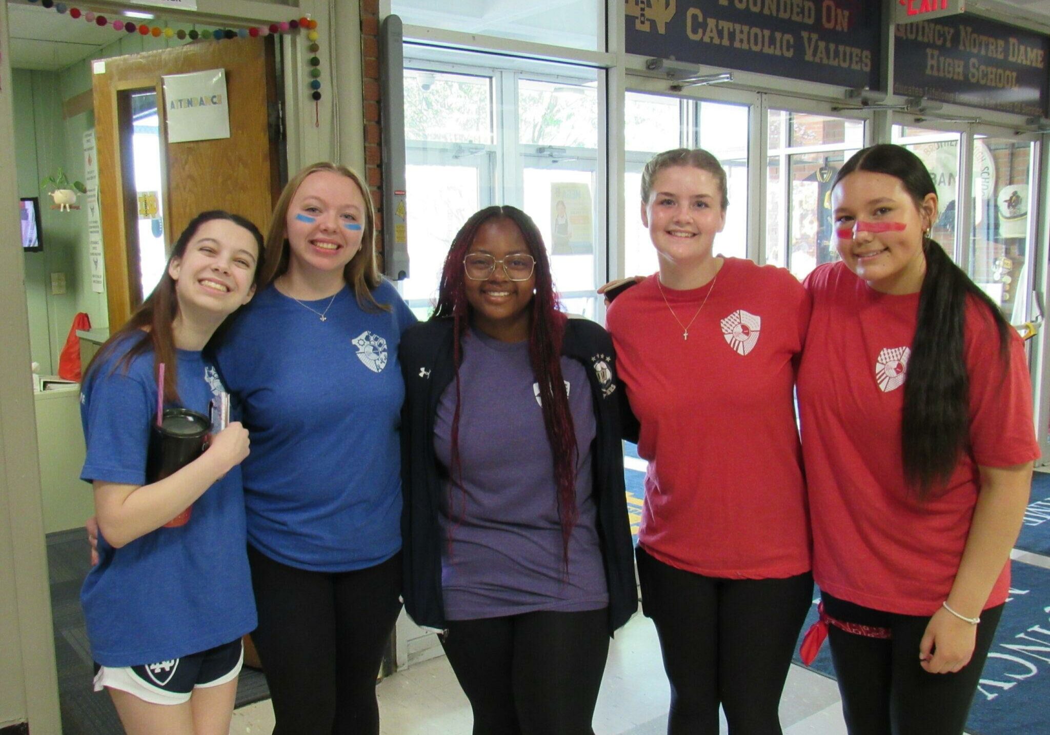 Five high school students smiling and posing together in a school hallway, wearing blue and red spirit shirts with face paint, under banners reading 'Founded on Catholic Values' and 'Quincy Notre Dame High School.