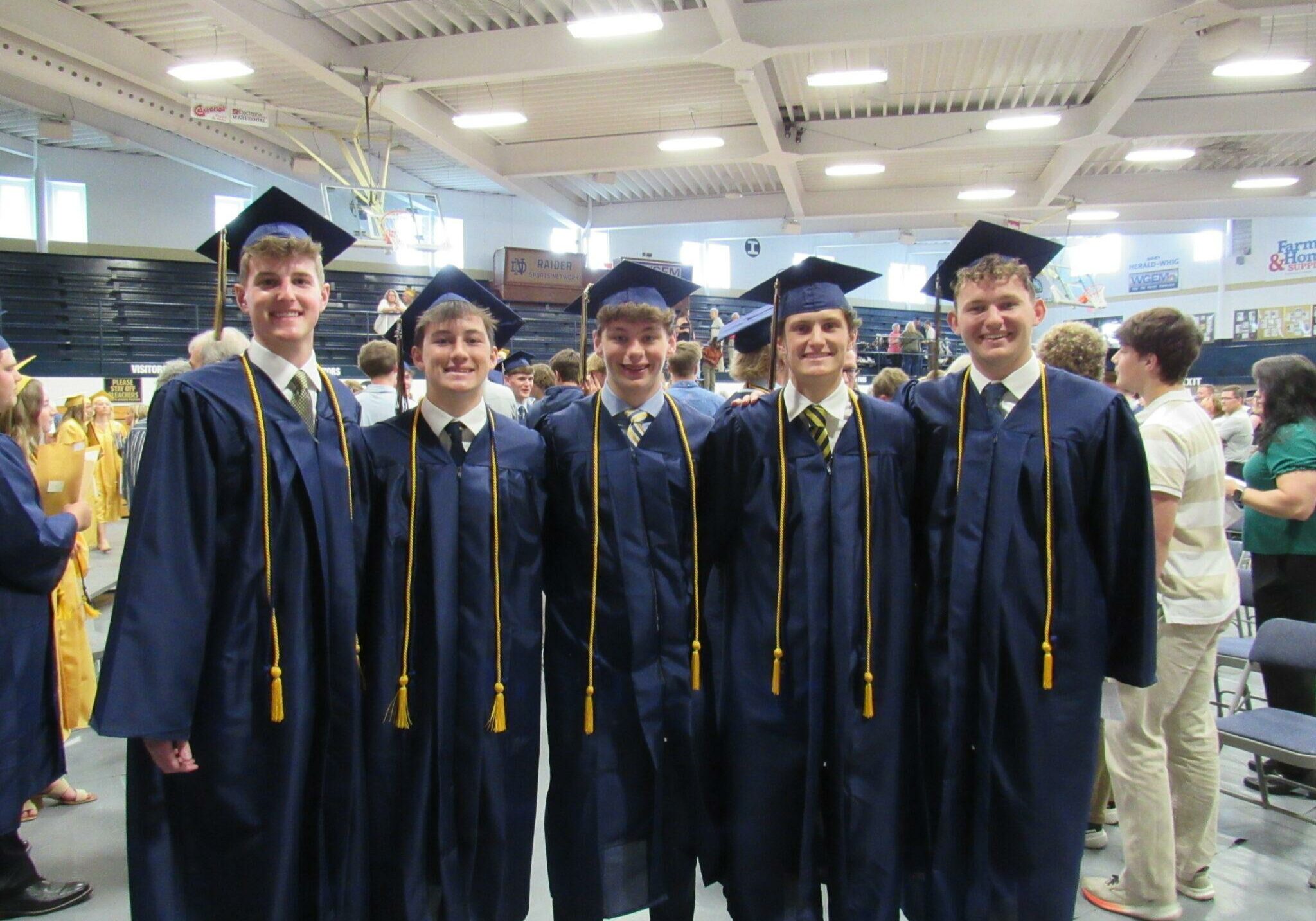 Five Quincy Notre Dame High School graduates in navy blue caps and gowns smiling during indoor graduation ceremony.