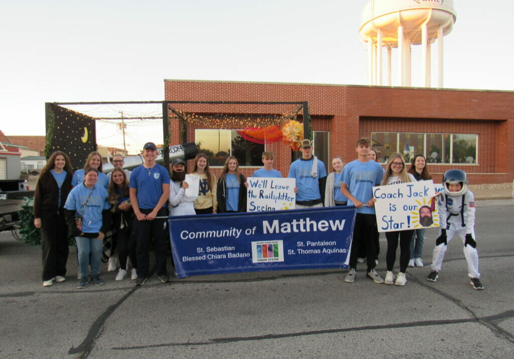 The QND Community of Matthew students stand in front of their parade float and hold a community banner.